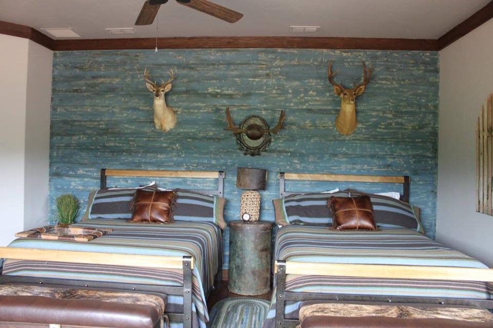Texas hunting lodge bedroom with distressed teal barn-wood accent wall, deer mounts, iron pipe bed frames, and striped bedding — Tincher's Custom Homes